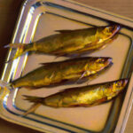 Three golden-yellow fish, a creation by the Duluth MN artist, are placed on a reflective metal tray against a brown table surface. The fish, arranged side by side, boast glistening scales and visible fins. The lighting gives them a shiny, lustrous appearance.