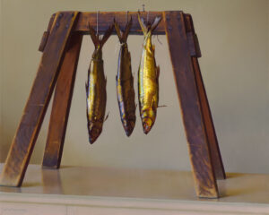 Three fish, captured by a Duluth MN artist, hang upside down by their tails from a wooden stand resembling a small A-frame. This artistic setup rests on a light-colored surface against a plain beige background.