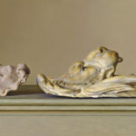 Two classical-style sculptures rest on a surface. On the left, a serene face with curly hair is depicted; on the right, an intricate relief showcases multiple faces intertwined in a dynamic composition. Created by a talented Duluth MN artist, both pieces are set against a neutral background.