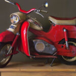 A vintage red and beige scooter displayed on a wooden platform against a dark background, with a rounded headlight and wide handlebars.