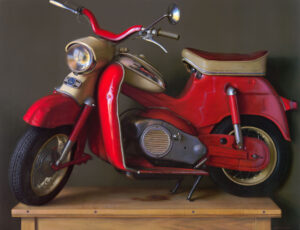 A vintage red and beige scooter displayed on a wooden platform against a dark background, with a rounded headlight and wide handlebars.