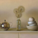 A still life scene on a beige table, crafted by a Duluth MN artist, features a shiny silver teapot, a glass bottle with dried dandelions, and a stack of white bowls with a ball of twine on top. The background is softly lit, creating a calm atmosphere.