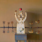 A creation by a Duluth MN artist, this sculpture stands with arms raised, topped with a red apple. A rod adorned with stars graces the left, while whimsical duck figures charm the right against a warm beige backdrop.