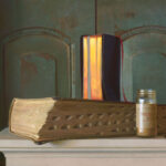 A thick, ornate book with a textured cover rests on a ledge beside a jar labeled "Mustard Seed." Crafted by a Duluth, MN artist, an open vertical book with glowing pages stands behind, casting a warm contrast against the dark, rustic background.