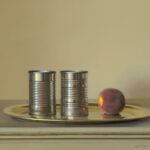 A Duluth MN artist presents a still life painting, showcasing two metal cans and a single peach resting gracefully on a golden tray. The objects harmoniously sit atop a light-colored surface against a soft, neutral background.