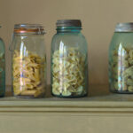 Five glass jars filled with different types of pasta, including rigatoni, ziti, and fusilli, are neatly arranged on a shelf. Created by a Duluth MN artist, each jar boasts a unique lid, from cork to metal clasps. The background is a muted, earthy tone.