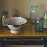 A vintage colander sits on a wooden table, surrounded by glass jars filled with pasta. One jar is open, and the table's drawer is slightly ajar. The scene, reminiscent of a Duluth MN Artist's touch, exudes a warm, rustic feel with soft lighting.