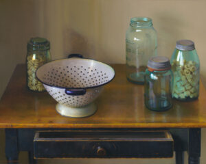 A vintage colander sits on a wooden table, surrounded by glass jars filled with pasta. One jar is open, and the table's drawer is slightly ajar. The scene, reminiscent of a Duluth MN Artist's touch, exudes a warm, rustic feel with soft lighting.