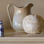 A Duluth MN artist captures a still life scene with a ceramic pitcher alongside a fabric-wrapped ball. To the left, a bottle adorned with blue and white designs adds charm. The objects rest on a light-colored surface against a neutral backdrop, showcasing the artist's keen eye for detail.