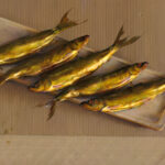 A realistic painting by a Duluth, MN artist features five golden fish arranged on a rectangular platter against a textured beige background. The diagonal placement of the fish creates a sense of harmony and visual interest in the composition.
