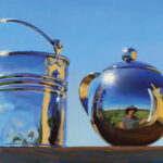 A reflective silver teapot crafted by a Duluth MN artist and a metal bucket sit on a wooden surface under a clear blue sky. The teapot's surface captures the distorted reflection of the landscape and a person taking the photo.