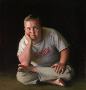 A person wearing a Red Sox t-shirt sits cross-legged on the floor, chin resting on hand, exuding a thoughtful demeanor reminiscent of a Duluth MN artist. The dark background artfully highlights their contemplative presence.