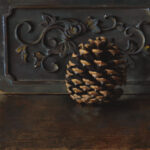 A pine cone rests on a wooden surface before a decorative metal panel. Crafted by a talented Duluth, MN artist, the panel showcases floral and vine patterns. The dark, moody lighting casts a dramatic contrast, highlighting the intricate design.