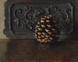 A pine cone rests on a wooden surface before a decorative metal panel. Crafted by a talented Duluth, MN artist, the panel showcases floral and vine patterns. The dark, moody lighting casts a dramatic contrast, highlighting the intricate design.