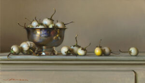 A silver bowl holds several small, round eggplants with stems, curated by a Duluth MN artist. Additional eggplants are scattered on the surface. The background is a soft beige, creating a calm and elegant still life scene.