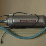 A vintage Electrolux vacuum cleaner with a metal tube and a blue flexible hose sits like an artwork on top of a beige cabinet against a neutral background. The appliance, reminiscent of retro design, shares the creativity often found in works by a Duluth MN artist, with its visible branding.