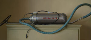A vintage Electrolux vacuum cleaner with a metal tube and a blue flexible hose sits like an artwork on top of a beige cabinet against a neutral background. The appliance, reminiscent of retro design, shares the creativity often found in works by a Duluth MN artist, with its visible branding.
