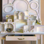 A still life scene by a Duluth MN artist features various white geometric foam structures arranged on a table. A bird, in motion, approaches a nest with blue eggs resting inside an open drawer, while two moths perch delicately on the foam.