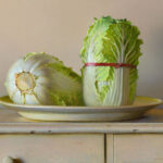 A realistic painting by a Duluth, MN artist depicts two heads of cabbage on a plate. One cabbage is upright, bound with a red band, while the other lies on its side, exposing the stem. The composition rests on a wooden surface against a neutral background.