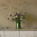 A bouquet of purple and white flowers with long grasses, arranged by a talented Duluth MN artist in a green glass vase, sits elegantly atop a cream-colored trunk against a beige background.