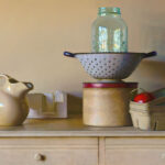 A vintage kitchen setup by a Duluth MN artist features a beige ceramic pitcher, a perforated metal colander with a glass jar, and a round tin box. A small rectangular basket with a tomato is artfully arranged on a light-colored wooden dresser.