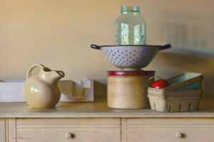 A vintage kitchen setup by a Duluth MN artist features a beige ceramic pitcher, a perforated metal colander with a glass jar, and a round tin box. A small rectangular basket with a tomato is artfully arranged on a light-colored wooden dresser.