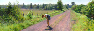 A Duluth MN artist in a gray dress bends down on a sunlit dirt road surrounded by lush green fields and trees. The scene is peaceful, with a clear blue sky framing the tranquil landscape.