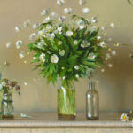 A Duluth, MN artist masterfully arranges four glass vases brimming with white flowers, including daisies, on a wooden surface against a light beige backdrop. Scattered blooms and petals enhance the scene with a natural touch.