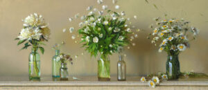 A Duluth, MN artist masterfully arranges four glass vases brimming with white flowers, including daisies, on a wooden surface against a light beige backdrop. Scattered blooms and petals enhance the scene with a natural touch.