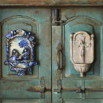 A rustic green door, crafted by a talented Duluth MN artist, features two ornate decorations: a blue and white ceramic plaque on the left, depicting a scene with figures, and a beige bas-relief on the right, showcasing a crucifix design.