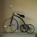A vintage blue tricycle with a worn and rusted frame, crafted by a Duluth MN artist, is displayed against a muted light brown background. This unique piece features two rear wheels, one large front wheel, a curved handlebar, and a slightly tilted seat.