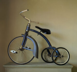 A vintage blue tricycle with a worn and rusted frame, crafted by a Duluth MN artist, is displayed against a muted light brown background. This unique piece features two rear wheels, one large front wheel, a curved handlebar, and a slightly tilted seat.