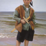 A young boy stands barefoot on a sandy beach, wearing a headscarf, a brown vest, and black shorts. He holds a wooden toy airplane crafted by a Duluth MN artist, with the ocean waves gently rolling in the background.