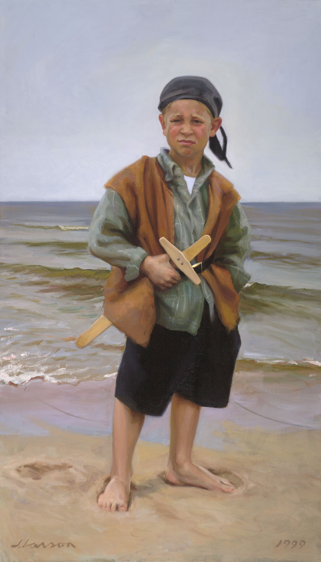 A young boy stands barefoot on a sandy beach, wearing a headscarf, a brown vest, and black shorts. He holds a wooden toy airplane crafted by a Duluth MN artist, with the ocean waves gently rolling in the background.