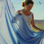 A Duluth MN artist stands in sunlight, holding a blue sheet on a clothesline. She wears a white tank top, and her hair is tied back. The background reveals a clear sky and open fields, capturing the serene beauty that inspires her art.