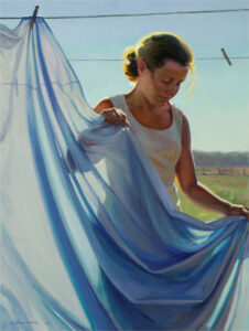 A Duluth MN artist stands in sunlight, holding a blue sheet on a clothesline. She wears a white tank top, and her hair is tied back. The background reveals a clear sky and open fields, capturing the serene beauty that inspires her art.