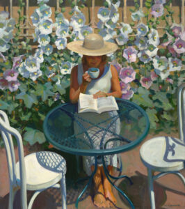A Duluth MN artist in a straw hat sits at a blue metal table, sipping from a cup and reading a book. She is surrounded by blooming white flowers on a sunny day, creating a serene garden atmosphere.