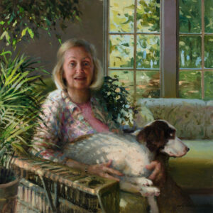 A Duluth MN artist with blonde hair sits in a sunlit room, surrounded by greenery, holding a large white and brown dog on her lap. Large windows in the background reveal a view of lush trees outside.
