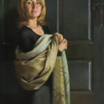 A woman with shoulder-length hair stands in front of a dark green door, holding an ornate, patterned fabric. She wears a black top and looks forward with a calm expression. Created by a Duluth MN artist, the painting embodies a classical and realistic style.