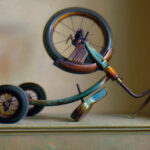 A vintage, rusted tricycle by a Duluth, MN artist is displayed upside down on a table, casting an intricate shadow on a plain background. The tricycle's worn paint and visible wear suggest age and use, with its handlebars and wheels prominently featured.