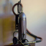 A vintage vacuum cleaner with a polished metal body and cloth hose, reminiscent of a Duluth MN artist's creation, stands on a wooden surface, casting a soft shadow against a light-colored wall.