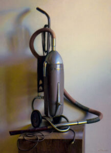A vintage vacuum cleaner with a polished metal body and cloth hose, reminiscent of a Duluth MN artist's creation, stands on a wooden surface, casting a soft shadow against a light-colored wall.