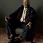 An elderly man, resembling a Duluth MN artist in his refined taste, sits in an armchair holding a glass. He's dressed in a formal suit and tie with a dark coat and red scarf, while his checkered socks add flair. The sophisticated ambiance is complemented by the muted background.