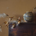 A Duluth MN artist captures a still life painting with a ceramic vase adorned with a blue pattern on a wooden surface. The vase cradles delicate, feathery white flowers and dried leaves, set against a warm, muted background, while seeds float gently in the air.