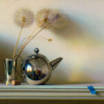 A still life by a Duluth MN artist showcases a metallic teapot and its reflection, beside a silver cup holding three dandelions. A small cork rests nearby, with a blue piece of tape at the edge against a soft, gradient background.