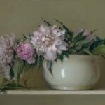 A white ceramic vase, crafted by a Duluth MN artist, holds a bouquet of pink and white peonies with leafy green stems, set against a muted olive-green backdrop. One peony lies gracefully beside the vase on a light-colored surface.