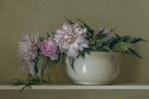 A white ceramic vase, crafted by a Duluth MN artist, holds a bouquet of pink and white peonies with leafy green stems, set against a muted olive-green backdrop. One peony lies gracefully beside the vase on a light-colored surface.