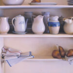 A kitchen shelf displays white and blue ceramic jugs, dishes, and mugs crafted by a Duluth MN artist. A neatly folded cloth rests on the lower shelf, beside three pears. The soft, natural lighting beautifully highlights the ceramics' smooth surfaces.