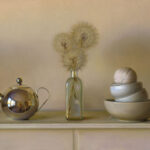 A polished silver teapot reflects its surroundings. Next to it, a glass bottle by a Duluth MN artist displays dried dandelions. Beside the bottle, three stacked bowls hold a ball of twine. All are arranged on a pale surface against a beige background.