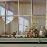 A rustic windowsill curated by a Duluth MN artist showcases a bird figurine, vibrant yellow flowers in a pot, a jar, and scattered dandelions. Light filters through the broken pane, casting serene reflections as a butterfly gently rests on the blossoms.